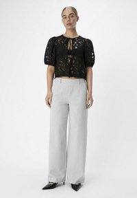 Haut en dentelle noire avec des manches bouffantes et un nœud à l'avant, associé à un pantalon large gris clair et des talons noirs pointus.