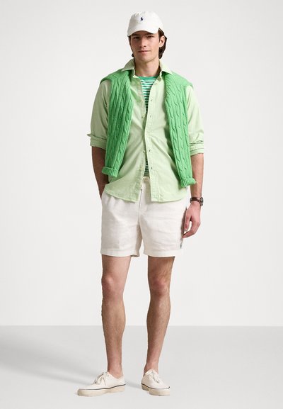 Chaleco de punto en verde claro sobre una camisa abotonada en verde claro, camiseta de rayas debajo, pantalones cortos blancos y zapatillas blancas. Gorro blanco en la cabeza.