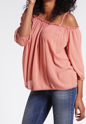 Vrouw draagt een losse, roze blouse met blote schouders en driekwart mouwen, gecombineerd met een blauwe jeans, poseert met één hand dicht bij haar gezicht.