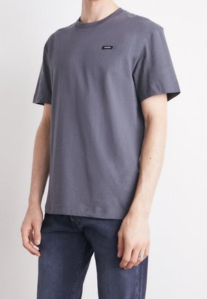 Mann trägt ein einfaches graues T-Shirt mit kurzen Ärmeln und kleinem Logo auf der Brust, kombiniert mit dunkelblauen Jeans und einem silbernen Ring an der linken Hand.