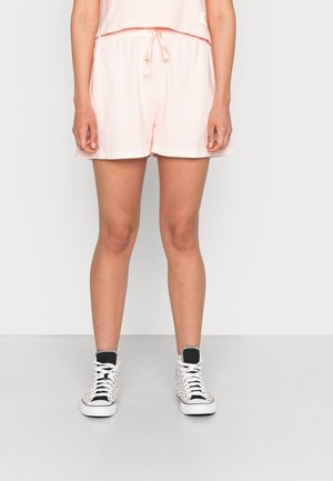 Osoba w jasnoróżowych szortach z wiązanym pasem i czarno-białych sneakersach za kostkę stoi na jasnym, jednolitym tle.