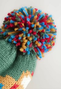 Grüne Strickmütze mit einem bunten Pompon, der rote, blaue, goldene und grüne Fäden enthält. Enthält geomtrische Muster in Beige und Weiß.