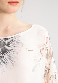 Blouse à imprimé floral, en tissu léger, avec un col rond et une coupe décontractée. Le design présente des illustrations de fleurs détaillées dans des couleurs douces.