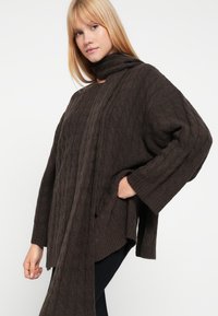 Stickad brun tröja med polokrage, som har ett texturerat kabelmönster och lösa ärmar. Inkluderar en lång, halsbandsliknande förlängning.