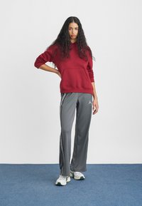 Kvinne med langt krøllet hår står med hånden på hoften iført en bordeaux hettegenser, grå joggebukser med striper, og hvite joggesko på blått gulv.