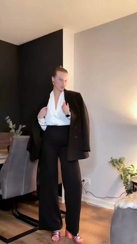 Blazer negro sobre una blusa blanca cruzada, combinada con pantalones negros de talle alto y sandalias de tacón rosas. Habitación con plantas y una silla.