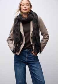 Beige getailleerde blazer over een donkerbruin sweater, gecombineerd met blauwe jeans. Een zwart en bruin patroon sjaal hangt om de hals.