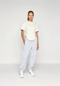 Šviesiai smėlio spalvos trumpomis rankovėmis marškinėliai su apvalia apačia, derinami su šviesiai pilkomis sportinėmis kelnėmis ir baltais sportbačiais su juodomis juostelėmis.