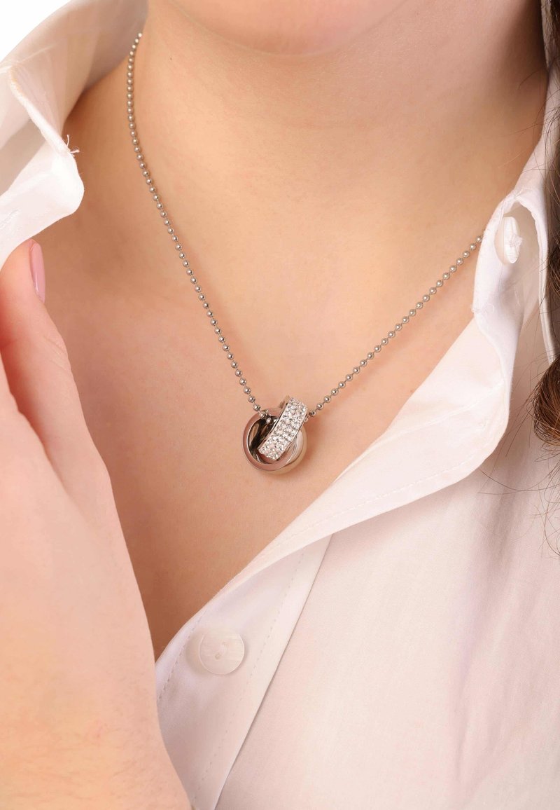 Zilveren ketting met in elkaar verstrengelde ringen, waarvan er één versierd is met heldere kristallen, hangend aan een balchain tegen een witte blouse.