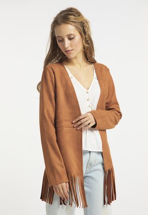 Vrouw draagt een bruine suède jas met franjes over een witte top met knopen en lichtblauwe jeans, kijkt naar beneden en raakt met één hand de jas aan.