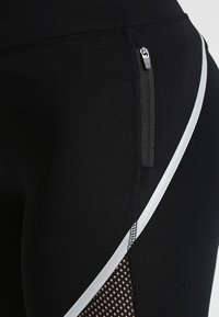 Detailní záběr černých sportovních legín s boční kapsou na zip, reflexním pruhem a síťovaným panelem na stehně.