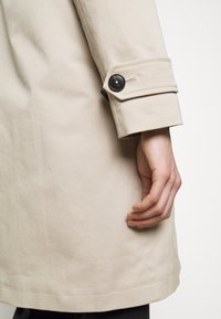 Beige trenchcoat met een gladde textuur, voorzien van een zwarte knoop en manchetdetail, getoond met een hand die rust op de stof.