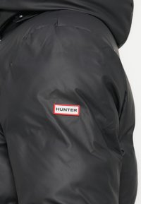 Melna pūdera jaka ar gludu tekstūru. Iezīmēta ar izteiktu sarkano un baltu "HUNTER" logo uz kreisā piedurknes.