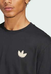 Svart bomull t-shirt med en vit broderad logotyp som liknar tre kronblad på vänster bröstområde, med en klassisk rund halsringning.