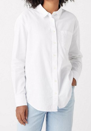 Žena v bílé košili s dlouhým rukávem a náprsní kapsou, v kombinaci s světle modrými džíny, ruka v kapse.