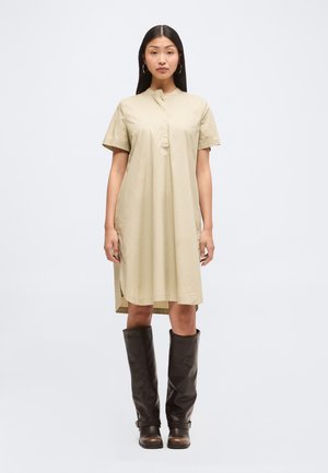 Femme aux longs cheveux foncés portant une robe-chemise beige boutonnée et des bottes en cuir brun foncé montant jusqu’aux genoux, debout sur un fond clair uni.