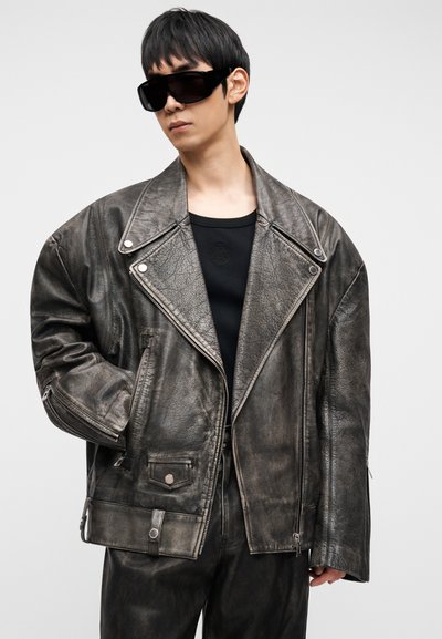 Suures hallis nahast mootorrattari jakk diagonaalse tõmblukuga, suured kraed, esietaskud ja hõbedased riistvaradetailid, kombineeritud musta topi ja päikeseprillidega.