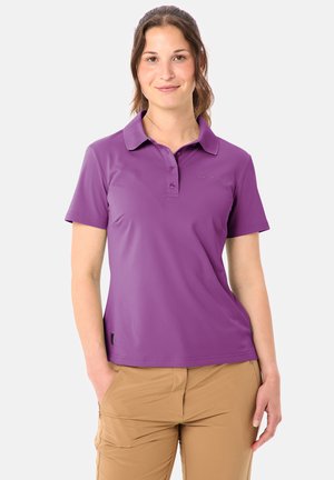 Femme portant un polo violet à manches courtes et un pantalon beige, debout avec une main dans la poche devant un fond uni.