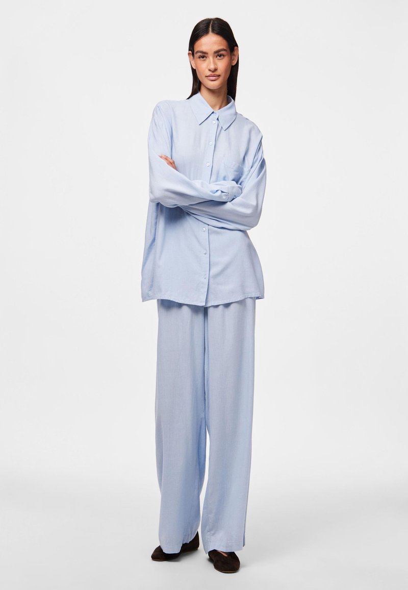 Camisa de botones de gran tamaño en azul claro con mangas largas y un bolsillo en el pecho, combinada con pantalones anchos a juego. Textura de tela suave.
