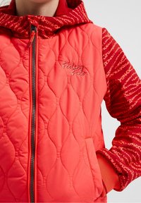 Czerwona pikowana kurtka z zamkiem z przodu, z wzorzystym kapturem i rękawami. Na piersi wyszywane hasło "Czuję się świetnie". Prążkowane mankiety.