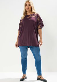 Bordeaux tunika med genomskinliga blommönstrade spetsärmar, avslappnad passform, matchad med blå skinny jeans och svarta ballerinaskor, mjukt tyg och subtil textur.