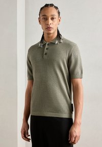 Koszulka polo z krótkim rękawem w oliwkowym kolorze, tekstura dzianinowa, zapięcie na pięć guzików, ściągacze przy rękawach i u dołu, akcent z boku, noszona z ciemnymi spodniami.