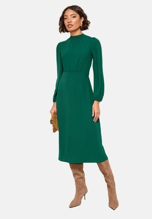 Grünes langärmliges Kleid mit hohem Kragen und gerafftem Bund. Verfügt über einen glatten Stoff und eine Midi-Länge, kombiniert mit beigen Stiefeln bis zum Oberschenkel.