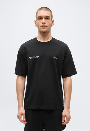 Lyhythiuksinen mies pukeutuneena mustaan ylisuurikokoiseen T-paitaan, jossa valkoinen teksti "MOTIVATIONAL QUOTE" ja "ICANWILL" rinnassa, seisoo valkoista taustaa vasten.