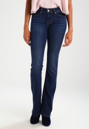 Femme portant un jean bootcut taille haute bleu foncé avec des coutures contrastantes, associé à des chaussures noires et un haut rose clair.