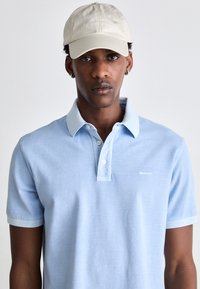 Camisa polo azul claro com mangas curtas, apresentando uma textura suave e um colarinho em contraste. Combinada com um boné bege com logotipo.