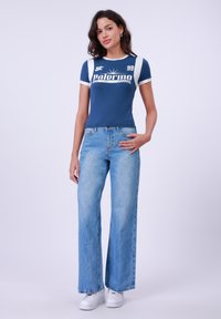 Top blu navy a maniche corte con dettagli bianchi e stampa grafica "Palermo 99", abbinato a jeans larghi azzurri e sneakers bianche.