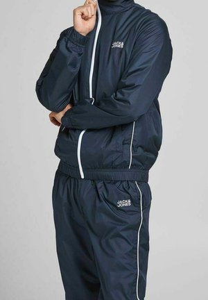 Survêtement marin comprenant une veste zippée et un pantalon à taille élastique, avec des détails en passepoil blanc et un petit logo sur la poitrine et la cuisse.