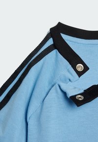T-shirt azzurra chiara con colletto nero, caratterizzata da strisce nere ai lati e bottoni a pressione. La texture in cotone morbido è ben visibile.