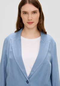 Blazer ajusté bleu clair en tissu texturé avec col à revers et fermeture à bouton bleu, porté sur un haut blanc.