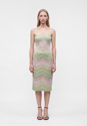 Frau trägt ein ärmelloses, knielanges Kleid mit pastellgrünen, rosa und silbernen Zickzackmustern und dünnen Schulterriemen, steht aufrecht.