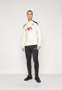 Męska długosleeve'owa koszulka sportowa w kolorze kremowym z czarnymi akcentami i czerwonym logo "AXA", zestawiona z czarnymi spodniami treningowymi z logo Nike.