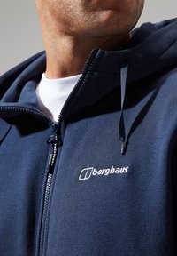 Navy zip-up hoodie featuring a textured fabric, adjustable drawstring hood, and white embroidered logo on the chest.