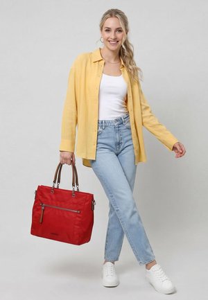 Mujer sonriente con cabello rubio, vestida con camisa amarilla, camiseta blanca, jeans azul claro y zapatillas blancas, sosteniendo un gran bolso rojo con asas marrones.