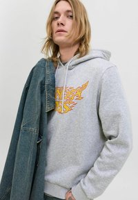 Sweat à capuche gris avec graphique de flamme orange, capuche à cordon, texture douce. Le modèle porte une veste en denim bleu sur son épaule sur un fond clair.