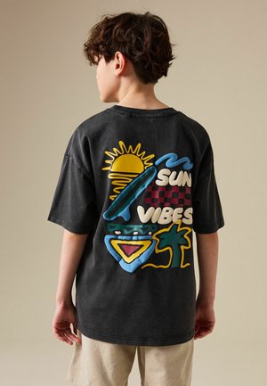 Kind mit lockigem Haar trägt ein schwarzes, übergroßes T-Shirt mit buntem Sonnen-, Palmen- und Surfbrett-Design und dem Schriftzug „Sun Vibes“ auf dem Rücken.