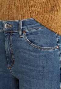 Blå denimjeans med medelhög midja, kopparknapp och sömdetaljer. Bärs med en texturerad senapsgul tröja.