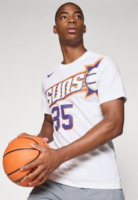 Canottiera da basket bianca a maniche corte con il logo "Suns" in viola e arancione, con il numero "35". Il giocatore tiene in mano un pallone da basket arancione.