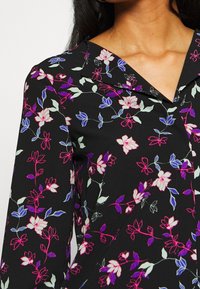 Chemise en tissu noir avec un col, ornée d'un motif floral en rose, violet et vert clair. Manches longues avec une texture lisse.