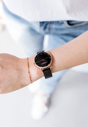 Montre en or rose avec un bracelet en maille noire, cadran rond et design minimaliste. Le cadran noir présente des index horaires blancs et un petit logo.