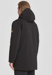 Zwarte jas met capuchon, vervaardigd van een gladde stof, in een getailleerde vorm, en met een subtiele logo patch op de mouw. Geverfd afgewerkt.