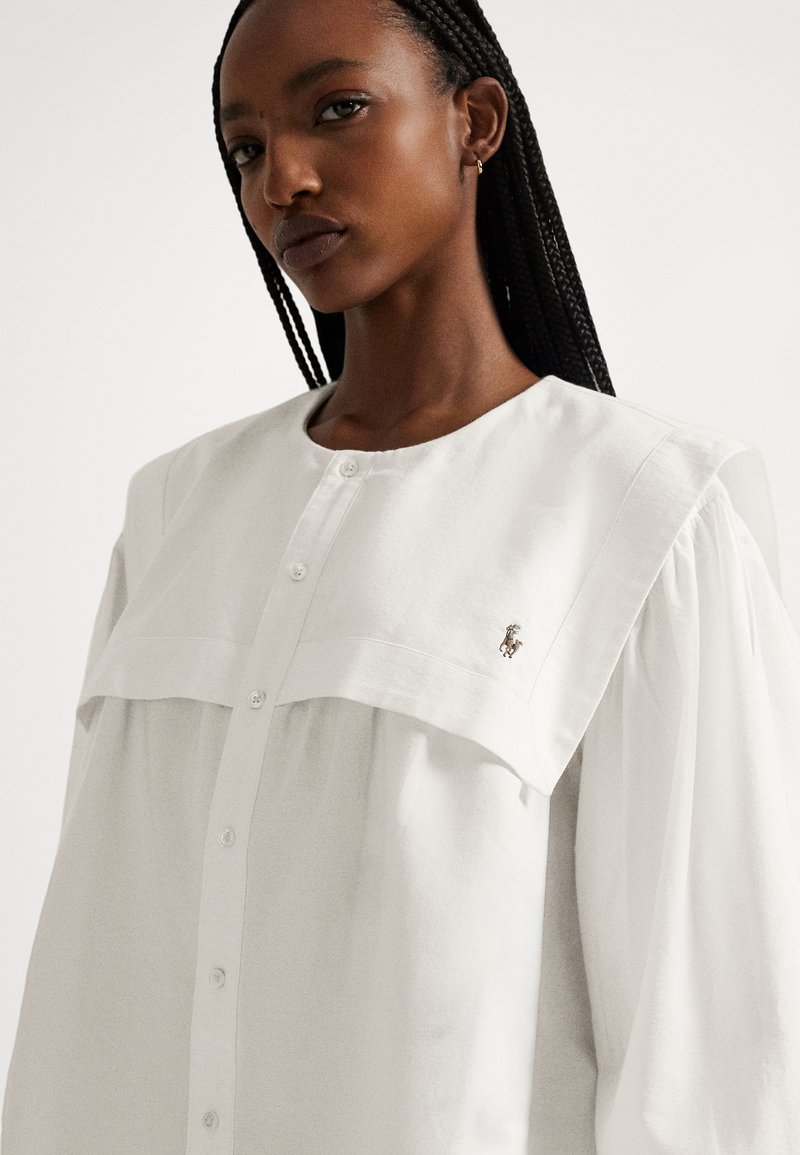 Vrouw met gevlochten haar, gekleed in een wit overhemd met knopen, brede kraag en een subtiel geborduurd logo op de borst, tegen een effen achtergrond.