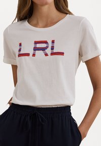 Balta medvilninė marškinė su suktais rankovėmis, dekoruota daugiaspalviu "LRL" siuvinėjimu raudona, mėlyna ir balta spalvomis. Kasdieniškas dizainas.