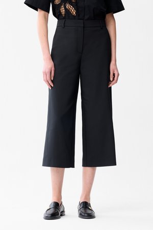 Persona che indossa pantaloni neri a gamba larga tagliati, mocassini neri e una maglia nera con un motivo in pizzo ritagliato visibile in vita.