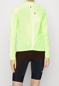 Veste de cyclisme légère jaune néon avec col montant, fermeture éclair et accents réfléchissants. Assortie à un short noir ajusté.
