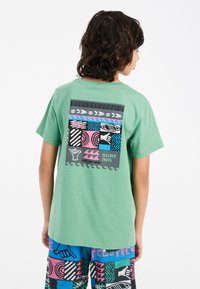 Camiseta verde de manga corta con un gráfico colorido en la parte posterior que presenta patrones inspirados en el surf, texto y diversas texturas.